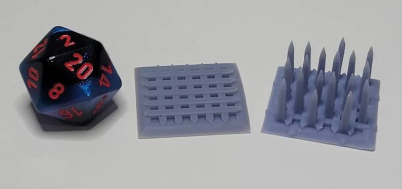 May include: Three miniature terrain pieces for tabletop games. A blue and black 20-sided die, a gray gridded square, and a gray square with a cluster of spikes.