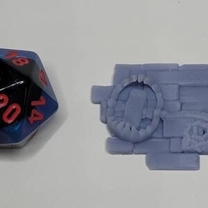 May include: A blue and black 20-sided die with red numbers, and a purple 3D printed miniature of a brick wall with a hole in it.