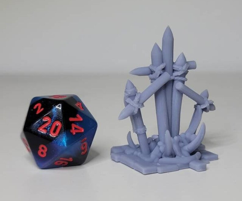 May include: A purple and black 20-sided die with red numbers sits next to a miniature model of a wooden stake trap. The trap is made of several wooden stakes bound together with rope and has a base of sharp, pointed spikes.