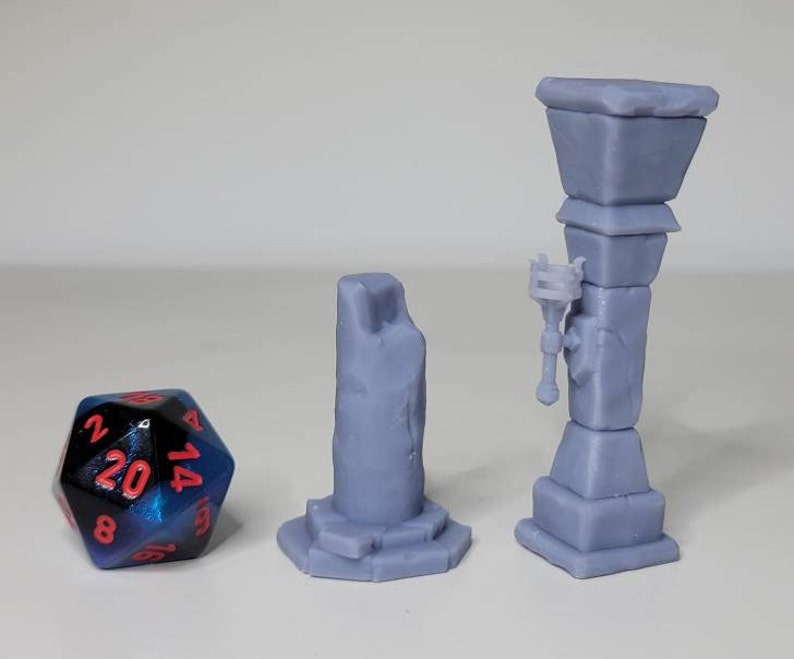 May include: Three 3D printed miniature stone pillars, one with a torch, for tabletop gaming or miniature display. The pillars are gray and have a rough texture. A blue and black 20-sided die is in the foreground.