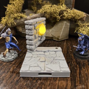 May include: A miniature fantasy scene featuring a gray stone wall with a glowing yellow light, a skeleton warrior with a sword, and a blue winged creature. The scene is set on a wooden surface, with moss in the background.