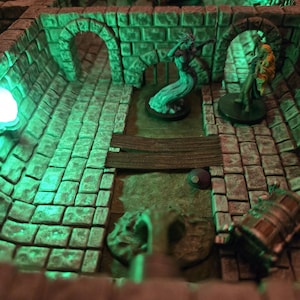 May include: A miniature dungeon diorama with green lighting. The scene includes stone walls, archways, and figurines of fantasy characters. A wooden bridge and a treasure chest are also visible, creating a detailed tabletop gaming environment.