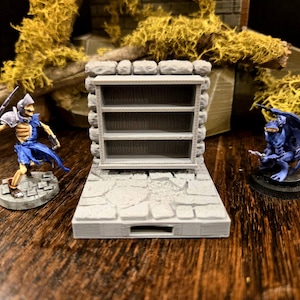 May include: A miniature gray stone bookshelf with three shelves is flanked by two fantasy figurines. A skeleton warrior in blue armor and a blue winged demon are positioned on a wooden surface, suggesting a tabletop game setting.