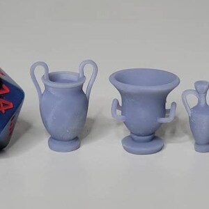 May include: Four miniature ceramic urns in various styles, including a tall urn with two handles, a shorter urn with a wide base, and two smaller urns with a narrow base. The urns are all a light gray color and are arranged on a white surface.