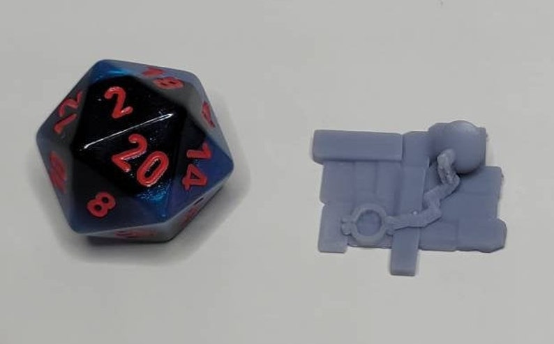 May include: A blue and black 20-sided die with red numbers and a small gray 3D printed miniature of a figure holding a chain.