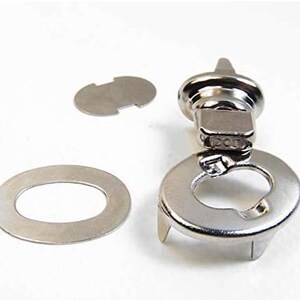 May include: A set of silver-toned metal purse hardware components. The set includes a clasp, an oval washer, and a small, irregularly shaped piece. The clasp has a rounded top and a rectangular base with a hinged mechanism.