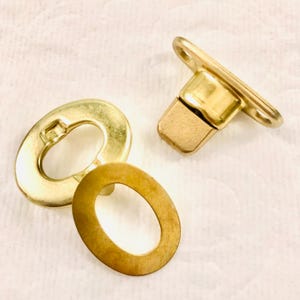 May include: A set of three gold-toned metal fasteners. The fasteners are oval-shaped with a hole in the center. The set includes two flat pieces and one piece with a raised center.