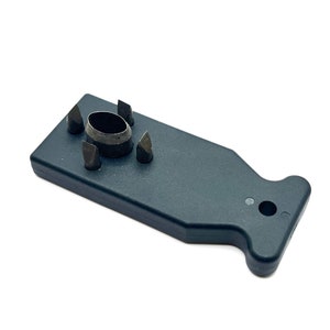 May include: A dark gray, T-shaped plastic tool featuring a central oval metal punch surrounded by four small, pointed metal spikes. The handle end includes a circular hole, and the overall design is flat with a matte finish for manual use.