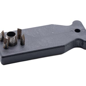 May include: A black plastic T-shaped handle tool featuring a central circular metal punch surrounded by four small, pointed metal attachments. The handle has a single circular hole near the top, and the metal components are fixed to the flat surface.