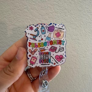 Puede incluir: Un portatarjetas colorido con la palabra "Phlebotomist" en letras de arcoíris. La insignia presenta ilustraciones de temática médica como un microscopio, jeringas y un monitor cardíaco. El portatarjetas tiene un cordón retráctil de plástico transparente y un clip metálico.