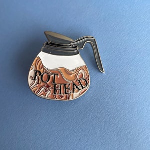 May include: A silver metal pin shaped like a coffee pot with a brown and white swirl design. The text "POT HEAD" is printed on the front of the pin.