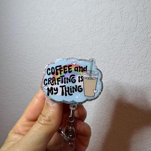 Könnte beinhalten: Ein Ausweis-Rollhalter mit dem Text "COFFEE and CRAFTING IS MY THING" in bunten Buchstaben. Der Ausweis-Rollhalter hat ein hellblaues Wolken-Design mit einer Kaffeetassen-Illustration. Der Rollhalter wird von einer Hand gehalten.