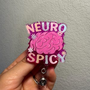 May include: A pink and purple badge reel with the words "NEURO SPICY" and a brain graphic. The badge reel has a clear plastic retractable cord and a metal clip.
