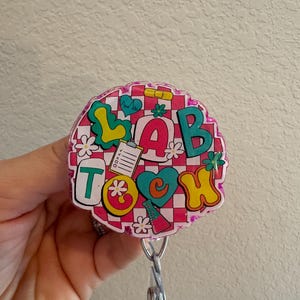 Puede incluir: Un portatarjetas redondo y colorido con las palabras "LAB TECH" en una fuente lúdica. El diseño incluye corazones, flores e iconos médicos sobre un fondo a cuadros rosa y blanco. El portatarjetas tiene un cordón retráctil de plástico transparente.