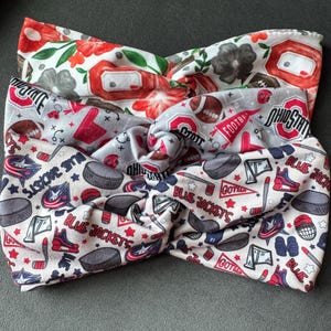Könnte beinhalten: Drei Stoffstirnbänder mit verdrehten Knotenmustern. Eines zeigt rote Blumen und graue Akzente, ein anderes Football- und Ohio State-Grafiken und das dritte Hockey-Muster mit dem Text "Blue Jackets".