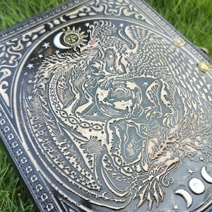 May include: A black leather journal with a detailed embossed design of a dragon and a phoenix intertwined around a globe. The design is in silver and gold, with stars and moons in the background.