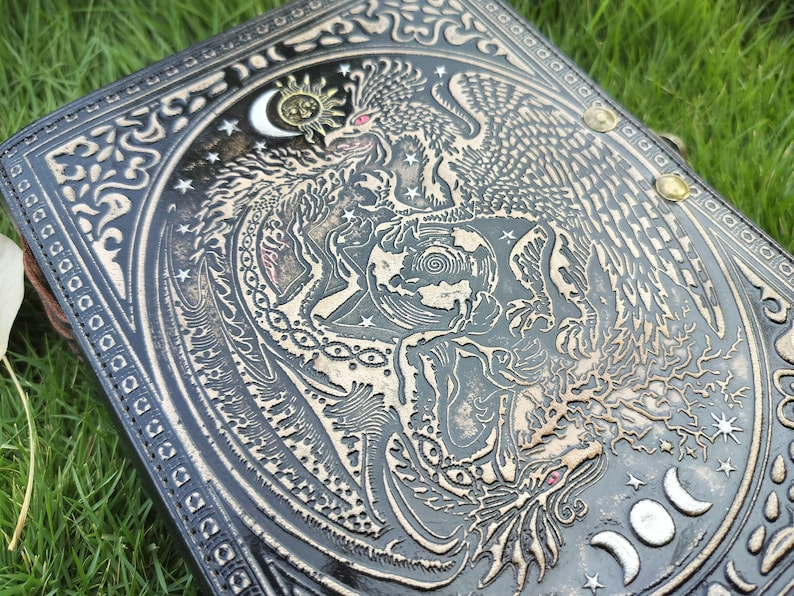 May include: A black leather journal with a detailed embossed design of a dragon and a phoenix intertwined around a globe. The design is in silver and gold, with stars and moons in the background.