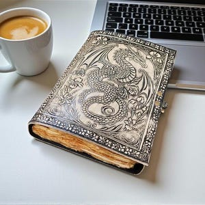 May include: A leather-bound journal with a detailed dragon design. The cover is cream-colored with a silver clasp. The journal is open, revealing aged paper. A white mug of coffee sits nearby, next to a laptop.