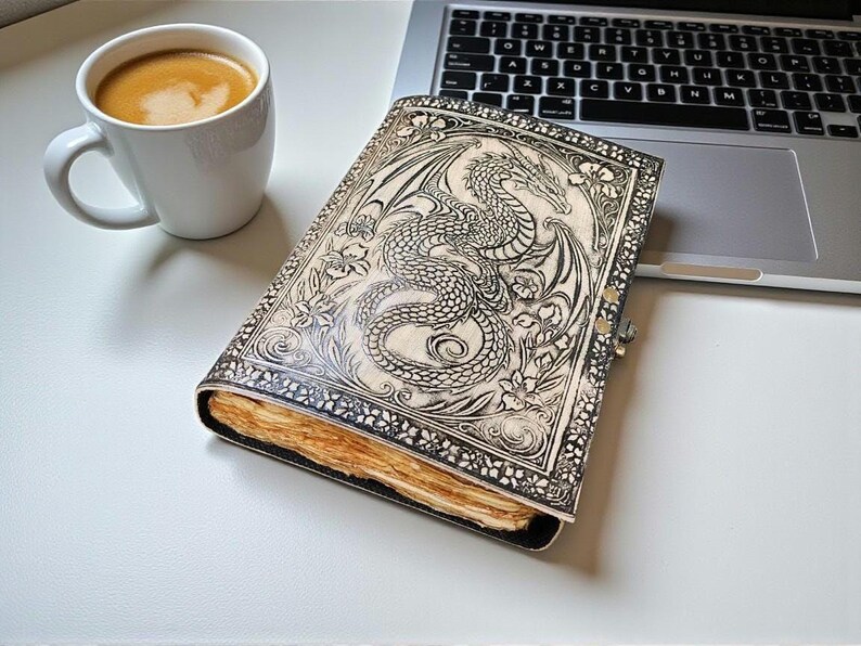 May include: A leather-bound journal with a detailed dragon design. The cover is cream-colored with a silver clasp. The journal is open, revealing aged paper. A white mug of coffee sits nearby, next to a laptop.