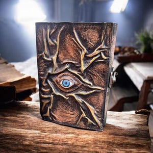 May include: A brown leather journal with an embossed design featuring an eye with a blue iris. The cover has a textured, aged appearance with intricate vein-like patterns. A metal clasp secures the journal.