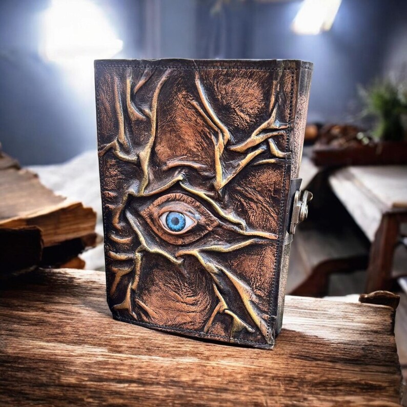 May include: A brown leather journal with an embossed design featuring an eye with a blue iris. The cover has a textured, aged appearance with intricate vein-like patterns. A metal clasp secures the journal.