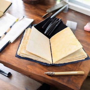 May include: An open, handmade journal with cream-colored pages sits on a wooden surface. A gold pen with ornate detailing rests nearby. Black pens and a small notebook are also visible, suggesting a workspace for writing or sketching.