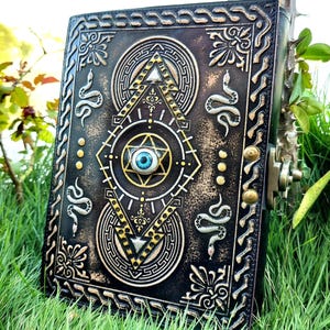Op de afbeelding: Een zwart lederen dagboek met een gouden reliëfdesign met een slang, een oog en geometrische vormen. Het dagboek heeft een messing sluiting.