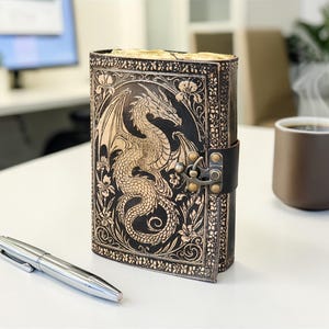 May include: A black leather journal with a dragon design embossed on the cover. The journal is closed with a leather strap and metal clasp. A silver pen and a brown mug of coffee are on the table.