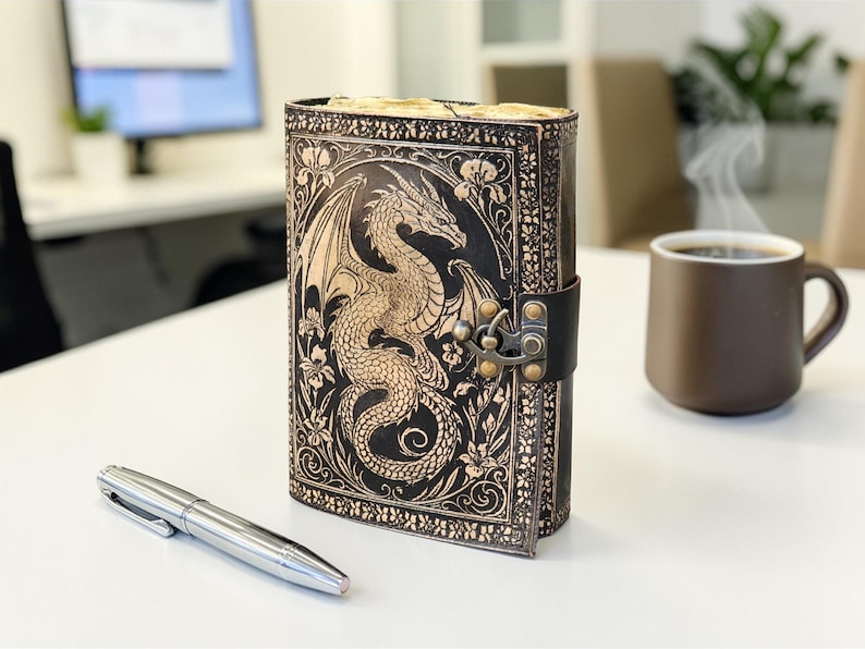 May include: A black leather journal with a dragon design embossed on the cover. The journal is closed with a leather strap and metal clasp. A silver pen and a brown mug of coffee are on the table.