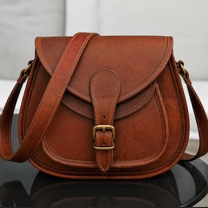 May include: A saddle-style crossbody bag in a rich, medium-brown tone. The bag features a rounded flap closure secured by a vertical strap with a brass-toned metal buckle. It includes an adjustable shoulder strap attached to brass-toned hardware.