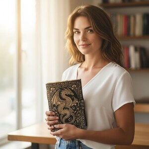 May include: A detailed, black leather journal with a gold dragon design and clasp. The journal is held by a person wearing a white top and blue jeans. The background includes a bookshelf and a wooden table.