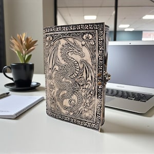 May include: A leather-bound journal with a detailed dragon design. The cover features an embossed dragon with intricate wing and scale details, surrounded by floral patterns. The journal is closed with a metal clasp.