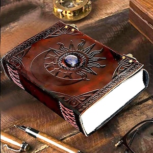 May include: A brown leather journal with a sun and moon design and a blue gemstone in the centre. The journal has a Celtic knot border and is closed.