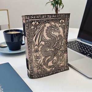 May include: A wooden journal with a detailed dragon carving on the cover. The dragon is surrounded by floral designs. The journal is standing upright on a white surface next to a laptop and a cup of coffee.