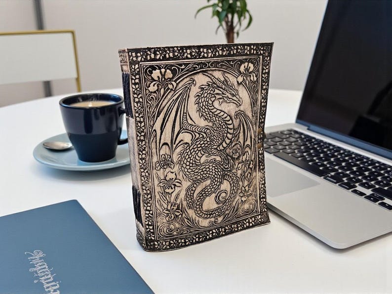May include: A wooden journal with a detailed dragon carving on the cover. The dragon is surrounded by floral designs. The journal is standing upright on a white surface next to a laptop and a cup of coffee.