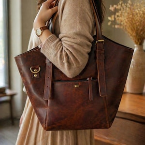 Pode incluir: Uma bolsa tote de couro marrom com um bolso frontal e alças ajustáveis. A bolsa tem ferragens de cor latão e um design estruturado. A bolsa está sendo carregada por uma pessoa vestindo um suéter e um vestido bege.