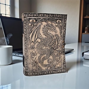May include: A wooden journal with a detailed dragon carving on the cover. The dragon is surrounded by floral designs. The journal is standing upright on a white surface, with a laptop and a coffee mug in the background.
