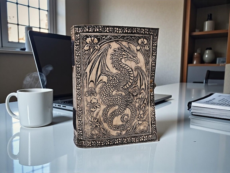 May include: A wooden journal with a detailed dragon carving on the cover. The dragon is surrounded by floral designs. The journal is standing upright on a white surface, with a laptop and a coffee mug in the background.
