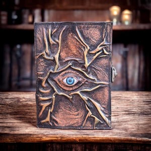 May include: A brown leather journal with an embossed design featuring an eye with a blue iris. The cover has a textured, weathered appearance with gold accents and a metal clasp. The journal is placed on a wooden surface.