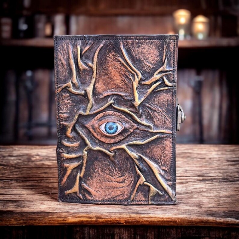 May include: A brown leather journal with an embossed design featuring an eye with a blue iris. The cover has a textured, weathered appearance with gold accents and a metal clasp. The journal is placed on a wooden surface.