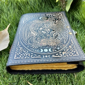 May include: A black leather journal with a gold embossed design of a dragon and a phoenix. The journal has a clasp closure and is decorated with a moon and stars.