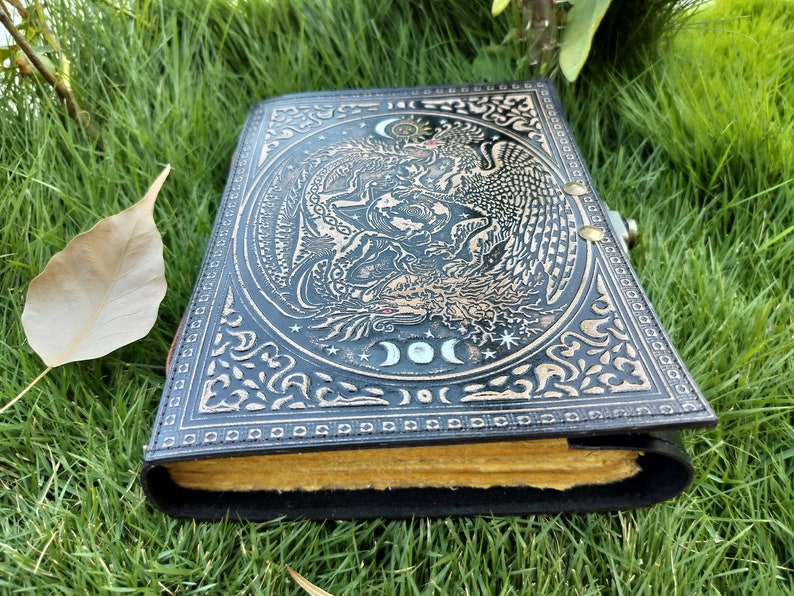 May include: A black leather journal with a gold embossed design of a dragon and a phoenix. The journal has a clasp closure and is decorated with a moon and stars.