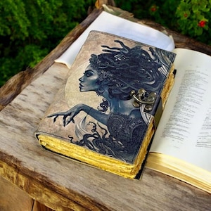 Puede incluir: Un diario encuadernado en cuero con una ilustración detallada de Medusa, la figura mitológica griega con serpientes por cabello, en la portada. El diario tiene un cierre de latón y un interior de papel amarillento y envejecido.