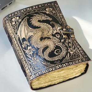 May include: A black leather journal with a detailed dragon design embossed on the cover. The dragon is tan, with intricate wing and scale details. The journal has a metal clasp and floral border. The pages are aged.