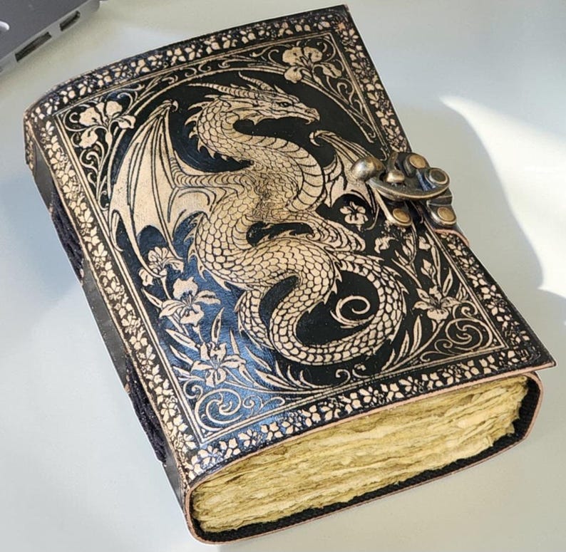 May include: A black leather journal with a detailed dragon design embossed on the cover. The dragon is tan, with intricate wing and scale details. The journal has a metal clasp and floral border. The pages are aged.