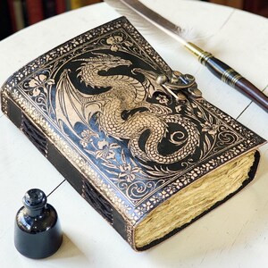 May include: A black leather journal with a dragon design embossed on the cover. The journal has a clasp closure and aged paper pages. A feather quill and inkwell are next to the journal.
