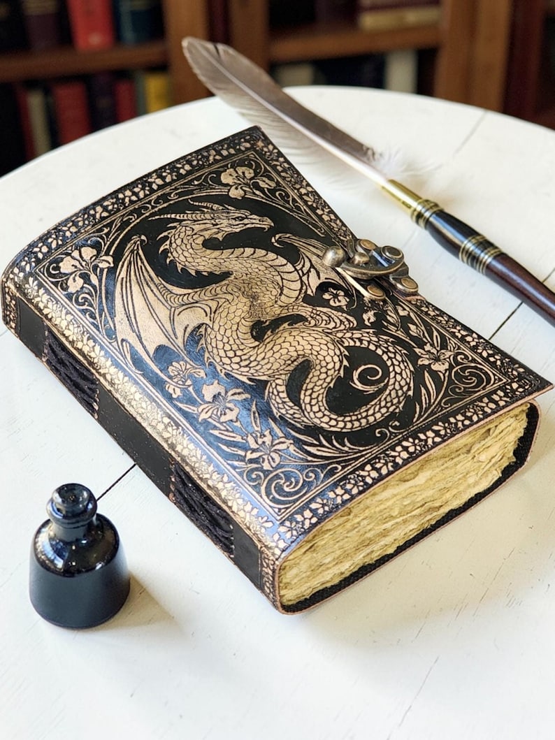 May include: A black leather journal with a dragon design embossed on the cover. The journal has a clasp closure and aged paper pages. A feather quill and inkwell are next to the journal.