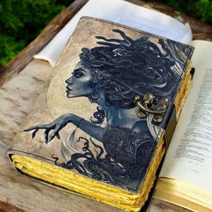 May include: A rectangular journal with a tan cover featuring a dark blue illustration of a figure with snakes for hair. The book includes a metallic latch closure on the side and has thick, textured, yellow-toned paper edges visible.