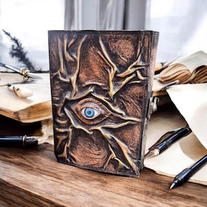 May include: A brown leather journal with a detailed, textured design featuring a blue eye in the center. The journal has a metal clasp and is placed on a wooden surface with pens and paper nearby.