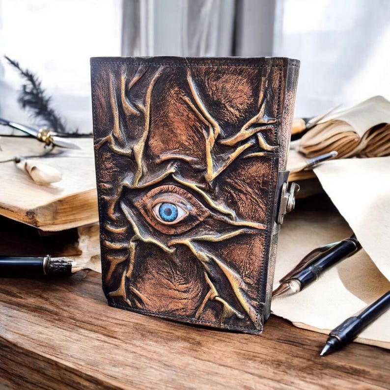May include: A brown leather journal with a detailed, textured design featuring a blue eye in the center. The journal has a metal clasp and is placed on a wooden surface with pens and paper nearby.